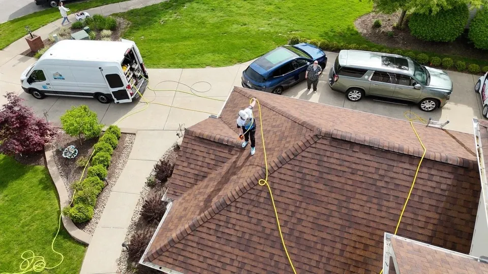 Aerial view of a technician soft washing a shingle roof, with the Patty's service van parked in the driveway below